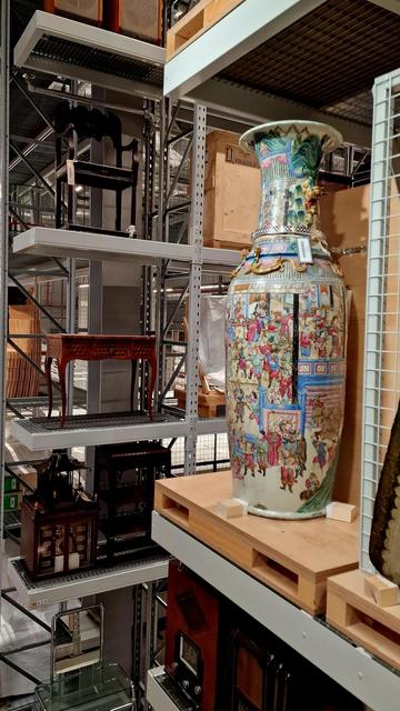 A Chinese vase painted in bold colours on a display shelf - five floors of shelves and items of furniture in the background