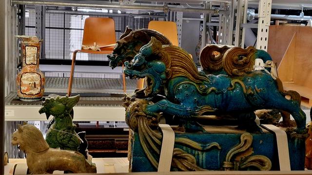 Two dragon-like beasts, painted in bold colours of turquoise and yellow on a shelf with two wooden chairs on shelves in the background
