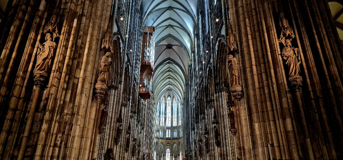 View into the central ship of the Kölner Dom with its high Gothic vaulted ceiling and intricate column design. The perspective shows an endless forest of column bundles in brown tones, while the ceiling glows in light greys and blues due to the stained glass windows bringing in light from the top