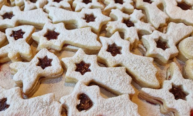 A whole tray of home made two layered biscuits in angel, snowflake and shooting star shapes - covered in icing sugar and with a star shaped cut out showing the layer of jam in between