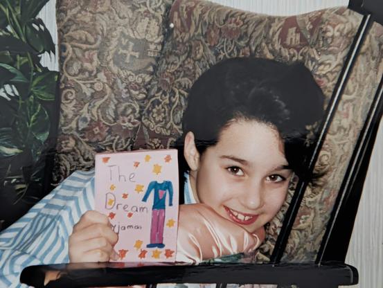 A young Kristie De Garis, dark-haired and grinning, propped on a patterned armchair, holding up a handmade picture book titled ‘The Dream Pyjamas,’ its cover hand-painted with a figure in blue and pink pyjamas and scattered autumn leaves.