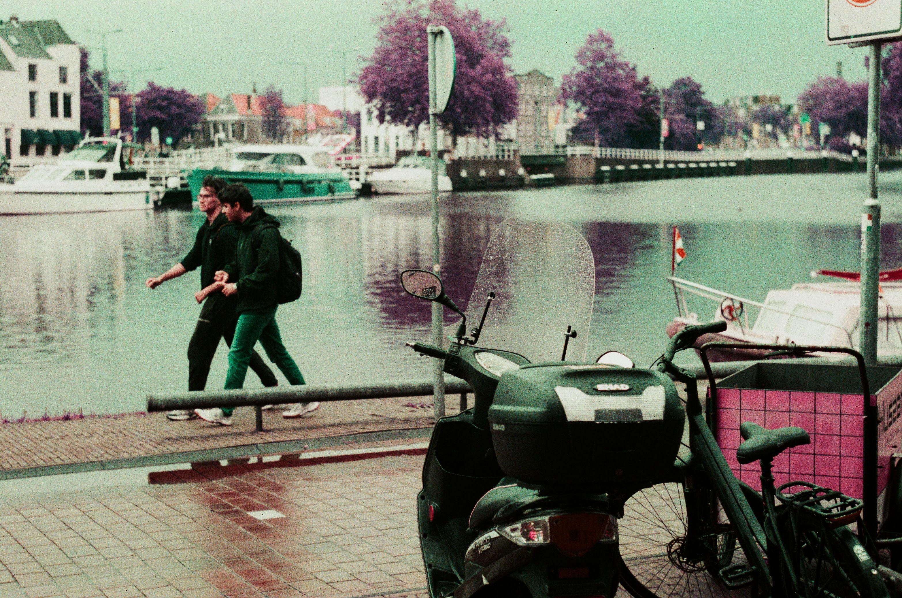 a purple-hued analog photograph of a wharf seen from across a path. the water is very cyan. in the foreground there are parked bikes and mopeds. two students are walking on the wharf in the left part of the frame. a boat lurks on the right side of the frame, in the water in the near background.