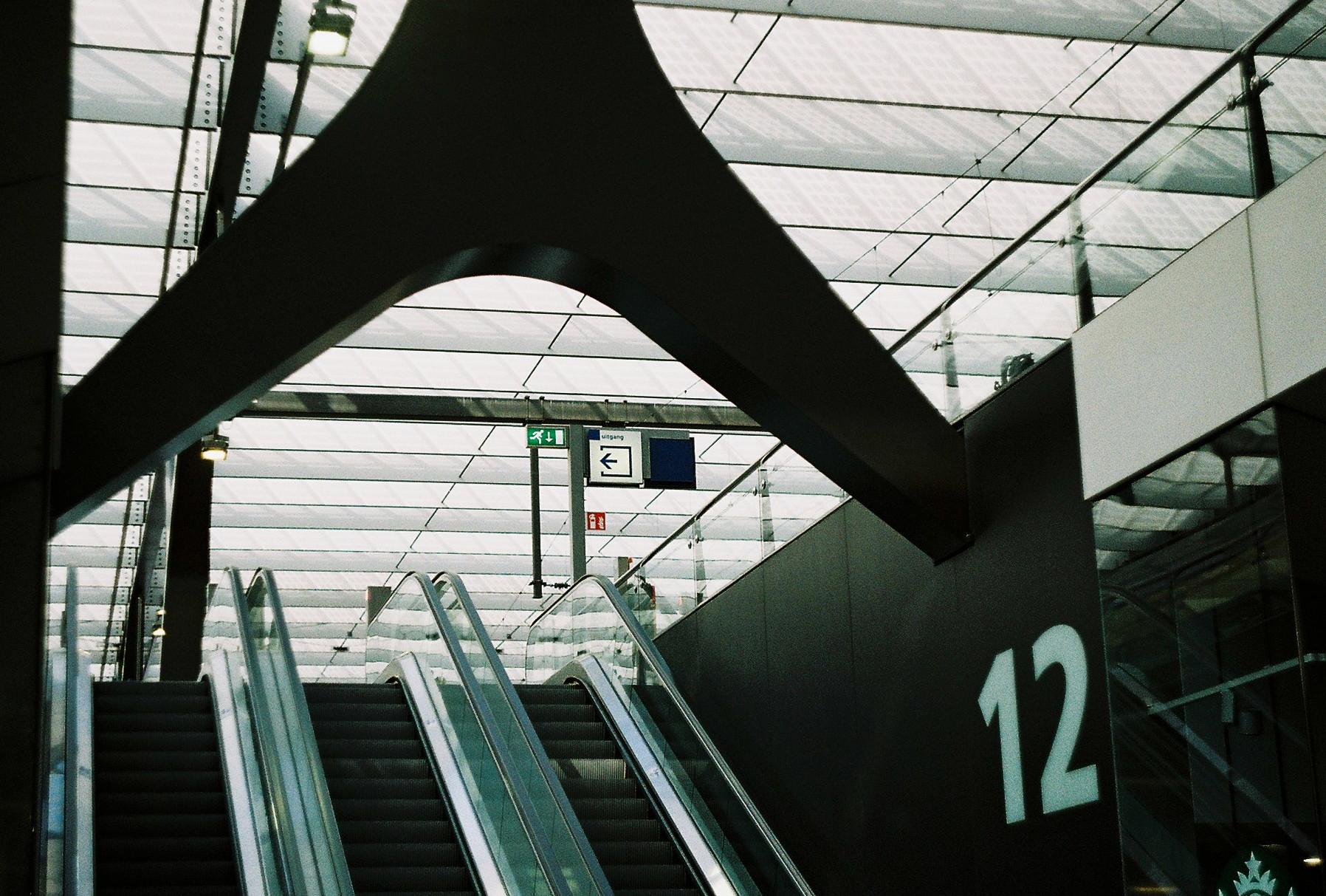 analog photo; escalators up to a platform at a very modern glass and steel train station. by the side of the escalators it says in very large font "12". the signage is clearly dutch. the ceiling shows a lot of the daylight and is very bright. blue, grey and white tones dominate the scene.