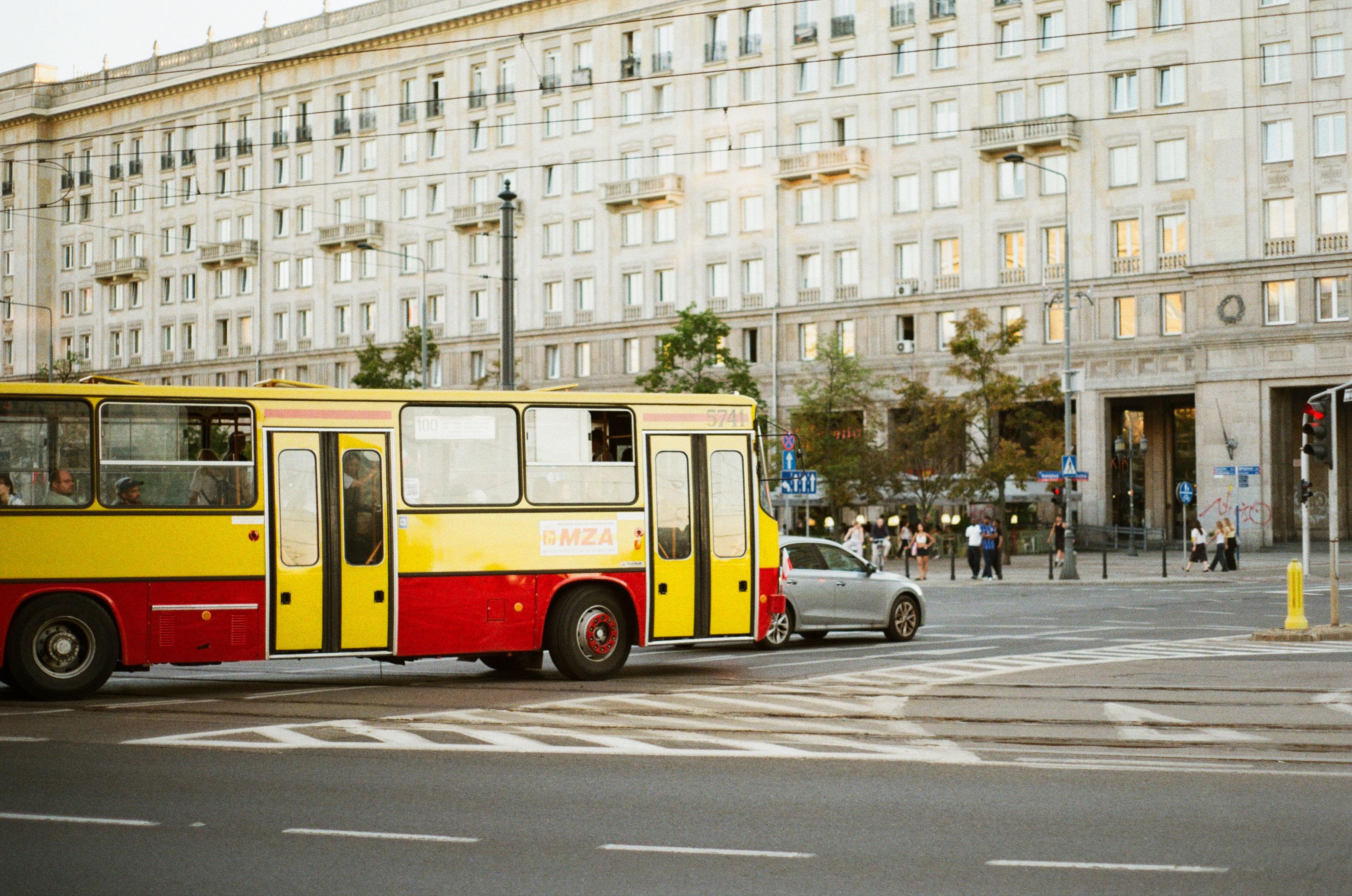 color film photo of a yellow and red bus turning on an intersection in warsaw. behind it is a massive concrete building with many neatly arranged windows.