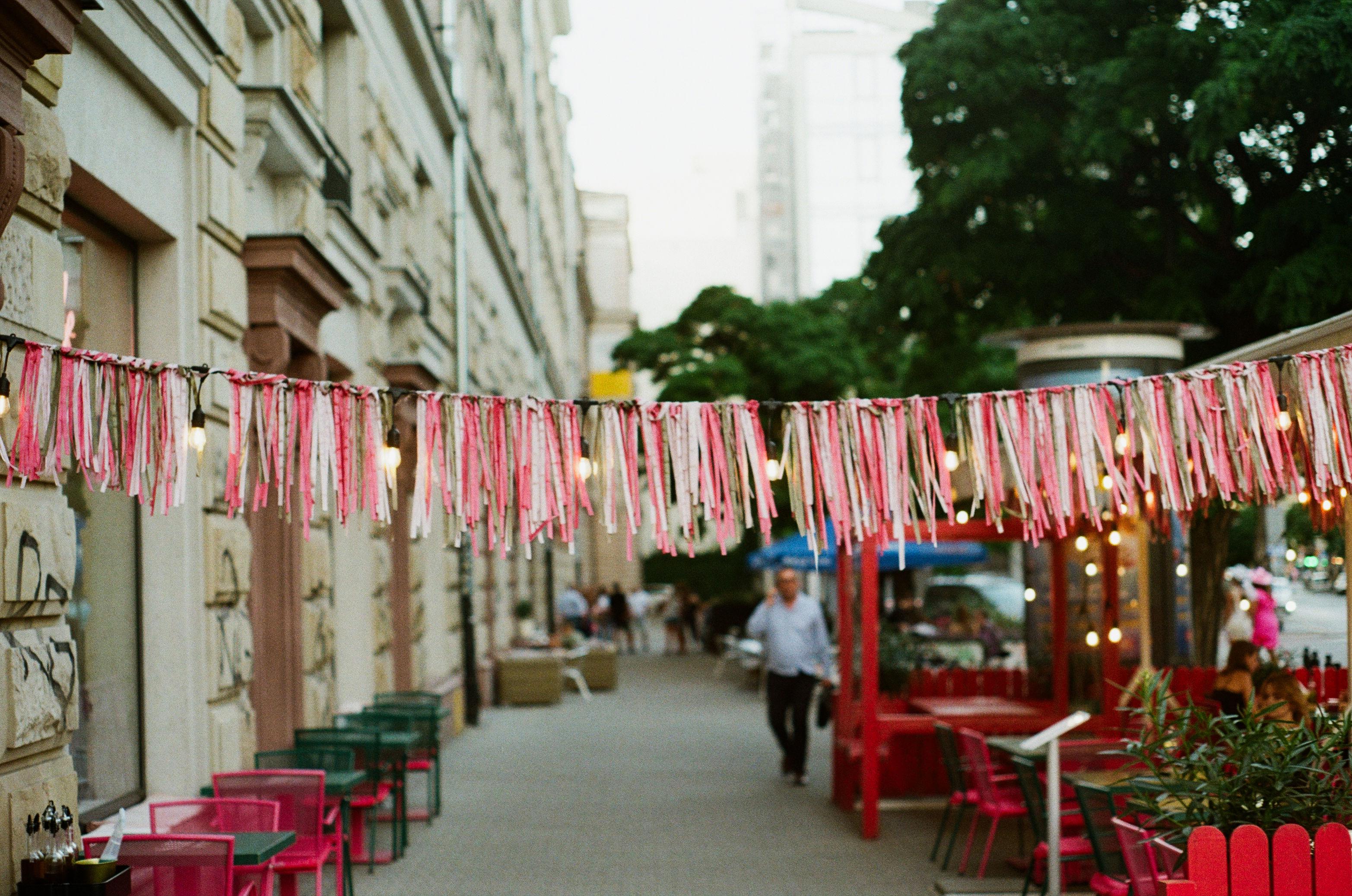 a color film photo of a cord with pink ribbons in various shades strung over the sidewalk. bright red chairs and tables in the background reveal that it's part of a restaurant's decor.