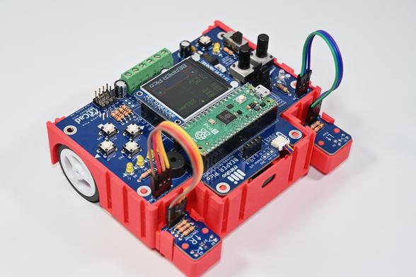 A blue BEAPER Pico circuit board controlled by a Raspberry Pi Pico microcontroller and with the optional LCD display is mounted in a red 3D-printed BEAPER Bot chassis. A white wheel is visible at the back (left), and two optical floor sensor modules are mounted at the front (right) connected by colourful extension cables. The black oval in-between the floor sensors is an optical time of flight ToF module for sensing distance.