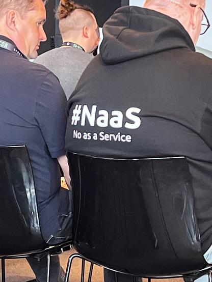 The image depicts a group of people, with a focus on the back of one individual wearing a black hoodie. The hoodie has the text "#NaaS" and "No as a Service" printed on it. This phrase is a play on the common technology term "as a Service" (e.g., Software as a Service, Infrastructure as a Service), humorously suggesting a service that offers "No" as a product. The setting appears to be an indoor event or conference, indicated by the presence of other attendees and what seems to be a lanyard on one person's neck. The overall tone of the image is likely humorous or satirical, aimed at those familiar with technology and service-oriented terms.