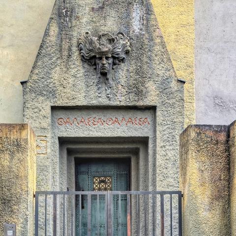 A ground-level view of a building entrance in Prague. A weathered stone facade features a centrally placed, muted teal double door with geometric metalwork. Above the doorway, carved in the stone, is a mask-like face with flowing hair, positioned beneath a Greek inscription in red lettering. A simple black metal railing spans the width of the entrance. The surrounding stone shows variations in color and texture, suggesting age and weathering.