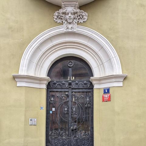 A detailed view of an Art Nouveau doorway in Prague. Ornate black metal gates with floral and geometric designs are set beneath a white arched frame. Above the arch, a stylized female face is carved in white stone, surrounded by floral motifs. To the right of the arch, a blue plaque displays the number "4," and below it, a red plaque reads "1162 PRAHA 1." The building's facade is a muted yellow.