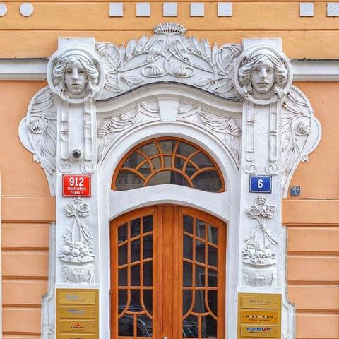 A detailed view of an ornate Art Nouveau doorway in Prague. The light brown wooden double doors are framed by a white, elaborately sculpted archway featuring stylized female faces on either side, surrounded by flowing floral and organic motifs. A red plaque to the left reads "912 STARÉ MĚSTO PRAHA 1," while a blue plaque on the right displays the number "6." Several small brass plaques with text are visible below the archway. The building's facade is a muted orange with subtle horizontal lines.