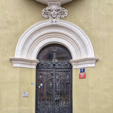 A detailed view of an Art Nouveau doorway in Prague. Ornate black metal gates with floral and geometric designs are set beneath a white arched frame. Above the arch, a stylized female face is carved in white stone, surrounded by floral motifs. To the right of the arch, a blue plaque displays the number "4," and below it, a red plaque reads "1162 PRAHA 1." The building's facade is a muted yellow.