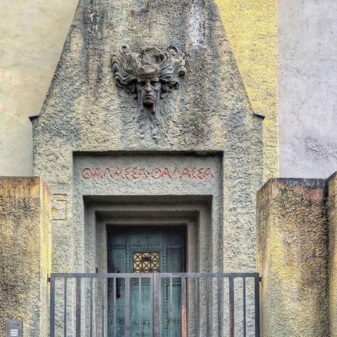A ground-level view of a building entrance in Prague. A weathered stone facade features a centrally placed, muted teal double door with geometric metalwork. Above the doorway, carved in the stone, is a mask-like face with flowing hair, positioned beneath a Greek inscription in red lettering. A simple black metal railing spans the width of the entrance. The surrounding stone shows variations in color and texture, suggesting age and weathering.