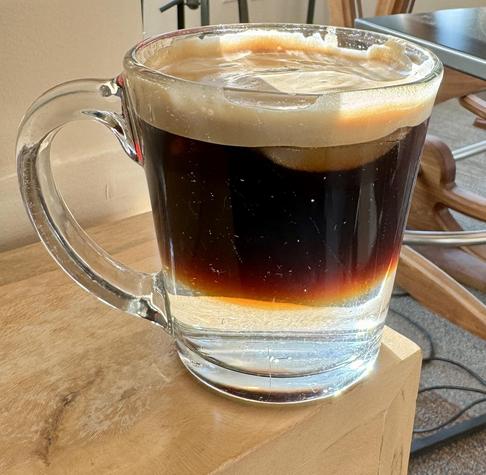 clear glass coffee mug with a clear layer of tonic water on the bottom slightly larger layer of espresso on top of that and a layer of foam on top with some ice floating through.