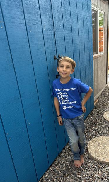 Young boy with blonde hair wearing a LibreTexts shirt and blue jeans smiling as he stands in front of a bright blue shed door