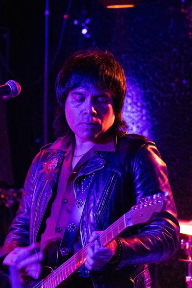 A dark-haired man wearing a leather jacket plays electric guitar, lit red and purple by stage lighting.