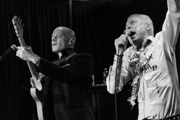 A bald man in a dark jacket plays electric guitar. Beside him, a white-haired man wearing a shirt printed with the words ‘fast clean cheap’ sings into a microphone.