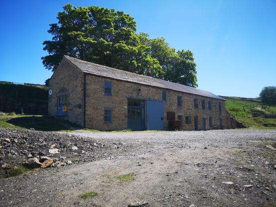 A long, two storey sandstone building has double doors, two single doors and several windows in its west elevation. Part of the roof is original stone, two thirds is metal sheet put on when this former derelict mine shop became a village hall, craft unit and upstairs bunkhouse. 3 sycamore trees stand behind against a blue sky.