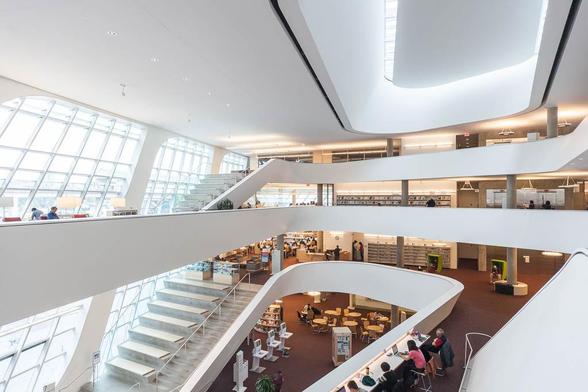 Interior of a contemporary library with curved white balconies, large windows allowing natural light, multiple floors with bookshelves, and people reading or working at tables on the lower level.