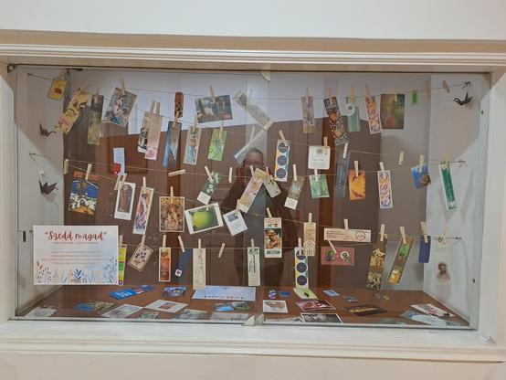 Photo of a window in the library wall with movable glass panels. Im the window there are strings to which several colorful bookmarks of all shapes and sizes are attacked with laundry clips. A sign in Hungarian says "pick your own"