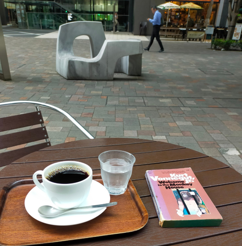 Photo is outside the cafe. An abstract white marble sculpture cum bench can be seen in the distance as well as a blurry Japanese man in blue dress shirt black dress pants looking at his phone while walking to the left. Further in the distance can be seen some sun blocking umbrellas for the outside seating at a restaurant. In the foreground is a plastic table of brown faux-wood. The tiny brown paperback book is on the right. A brown tray has a white mug & saucer of black coffee with a silver spoon in front. In the middle is a small clear tumbler of water
