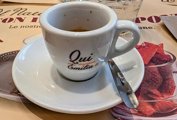A white espresso cup with the text "Qui si dispensa Emilia" sits on a matching saucer with a small metal spoon, placed on a table with a colorful placemat featuring images of desserts and Italian text. The cup contains remnants of espresso.