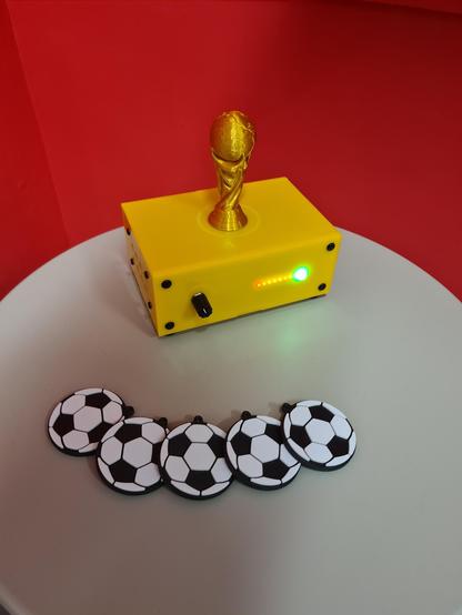 A small 3d printed replica of the World Cup sits atop a yellow Museum in a Box on a small circular white table. 

Also on the table sit 5 replica footballs.
