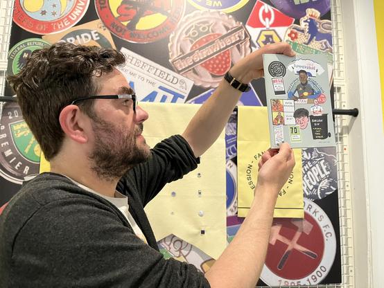 A man stands facing right looking at the edge of a fanzine so the front cover is visible to the camera.