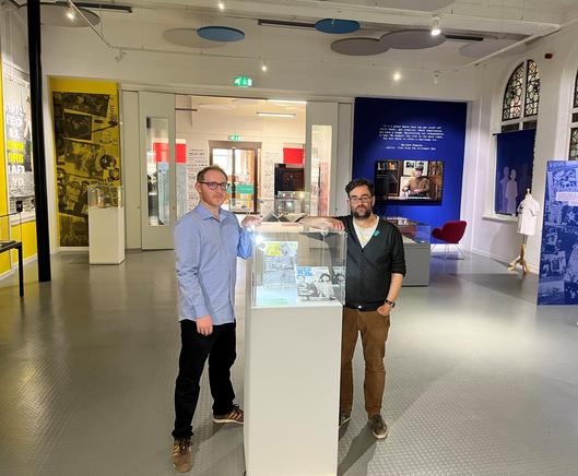 Two men stand in the middle of an exhibition room, to the sides we can see displays on the walls. The men lean on either side of a glass display case facing the camera.