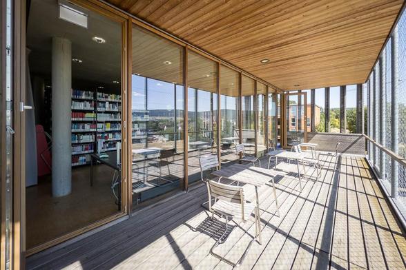 A wooden balcony with glass panels and netted safety rails overlooks a scenic view of greenery and buildings, which reflects in the windows. The space has several metal chairs and tables under a wooden canopy. The sky is clear, and sunlight casts shadows on the floor.