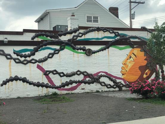 Mural on side of white wall of woman’s face, her long braids rendered in metal pieces attached to the wall