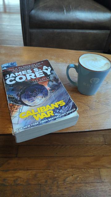 A table, on which is sitting the book "Caliban's War" by James S.A. Corey and a Violet latte (maple + blueberry) from Black River Roasters in Readington, NJ