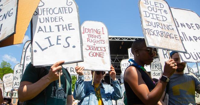 A photo of people holding up signs shaped like headstones and reading things like "Suffocated under all the lies" and "!dying! trans rights + services gone" and "bled out and died during miscarriage"