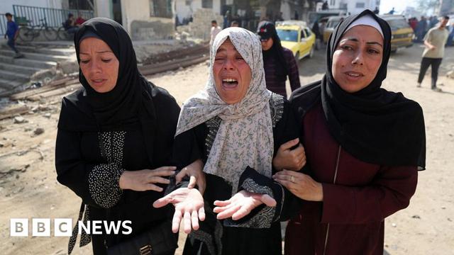 Three women linking arms with expressions of despair, with woman in the centre crying. Woman on left of image is wearing a black abaya with a black hijab, woman in centre, the same with a white printed hijab and the woman on the right is wearing a burgundy abaya with a black hijab and white bandana underneath it.