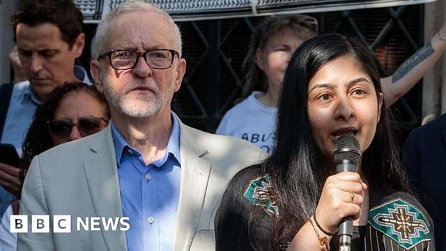 Jeremy Corbyn and Zarah Sultana stand among a group of people at a protest. Corbyn wears a light-colored blazer over a blue shirt. Sultana, dressed in a black outfit with colorful embroidery, holds a microphone.