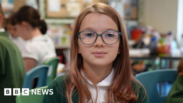 A school girl with long brown hair and blue rimmed glasses looks into the camera