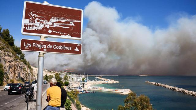 People look on near the Plage des Corbieres, in Marseille, southern France on 8 July 2025, as a smoke from a wildfire rages in the background. | © AFP - Clément Mahoudeau