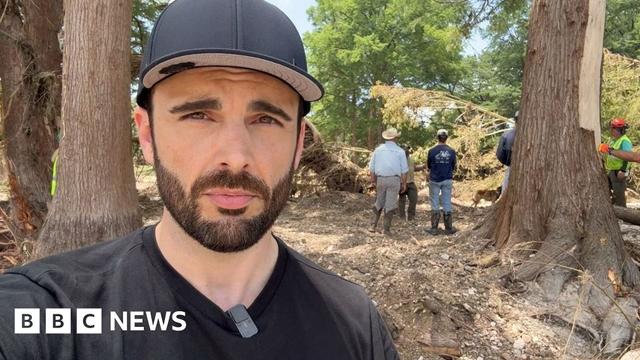 BBC reporter wearing a black cap with people in the background by trees
