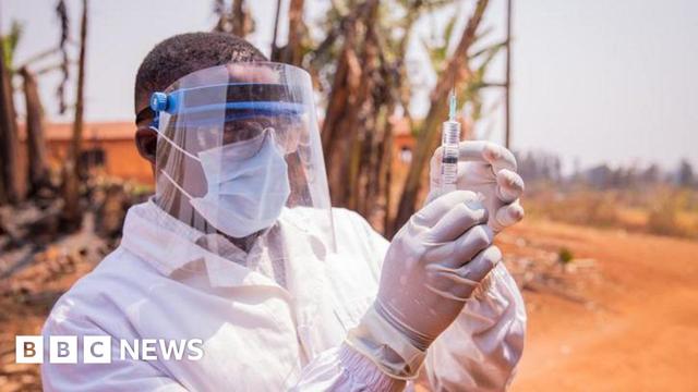 A medic in full protective face mask and white clothing, standing outside, holding up a syringe with gloved hands