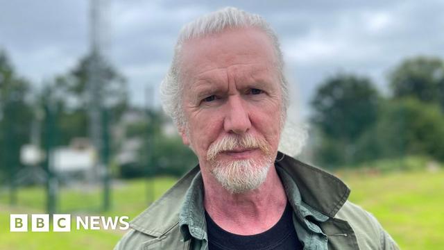 Shane Kelly, a man with white hair and beard, looks into the camera. He is wearing a green coat over a black t-shirt and a green shirt.