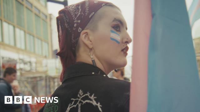 Mason is at a rally and looking over his shoulder. He is wearing make-up and a dark-red bandana. He has a blue, pink and white transgender Pride flag painted on his face.