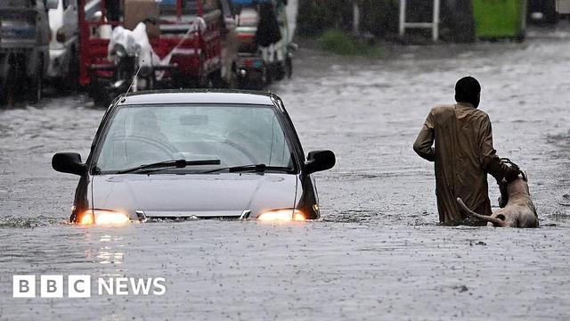 A man in a gray long-sleeved shirt inches along a flooded street with a white dog. He grabs its leash while a car wades through the same street during heavy monsoon rains in Rawalpindi, Pakistan on 17 July, 2025.