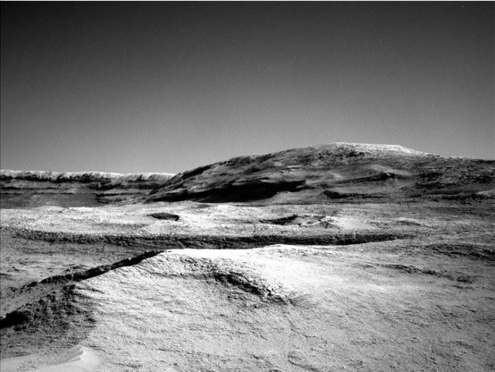 A grayscale photo from the Martian surface shows a landscape of undulating but mostly smooth terrain from the front of the image to a gently sloping hill that rises from the center of the image toward the upper right side of the frame. Beyond that, a straight ridge creates a horizon line along the left side of the image, disappearing from sight behind the hill.