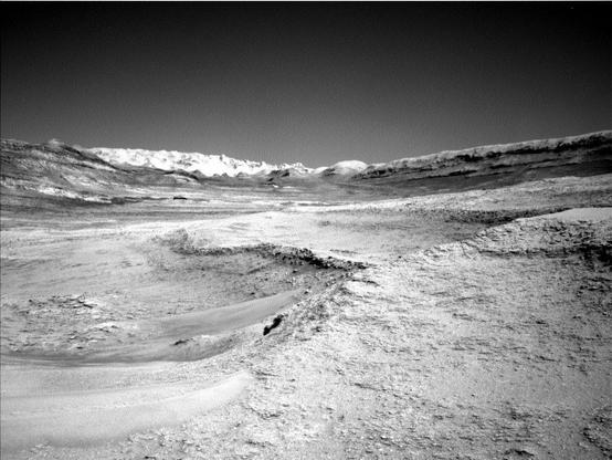A grayscale photo from the Martian surface shows a landscape of undulating but mostly smooth terrain, in light gray. A plateau on the right side of the foreground drops down toward the left about mid-frame, in a gentle curve. A ridge across the upper middle of the frame creates a V-shaped horizon line. Beyond that in the distance, filling the bottom of the V, a jagged line of rocks is brilliant white, looking like snow-capped mountains.