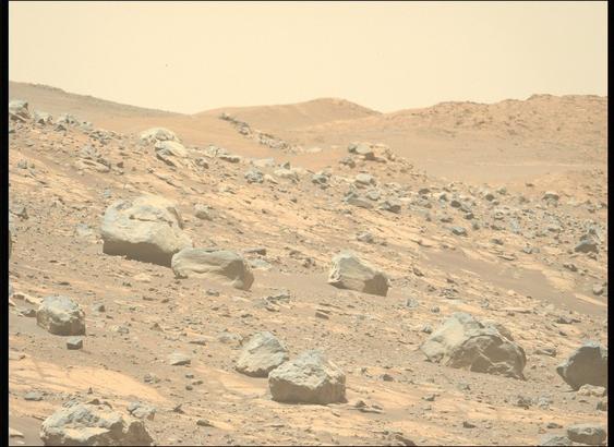 A color photo from the surface of Mars shows very rocky terrain in the foreground sloping downward from left to right. The ground ranges in shades from orange-tan to brown, while the rocks are mostly gray. A trio of hills rises up in the distance.