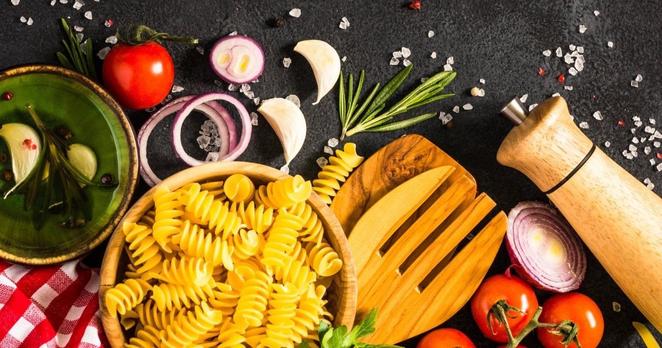 Eine Ansammlung von Lebensmittel mit Nudeln, Tomaten, Zwiebeln, Knoblauch Rosmarin und einigen groben Salzkörnern auf einem schieferdunklen Hintergrund.