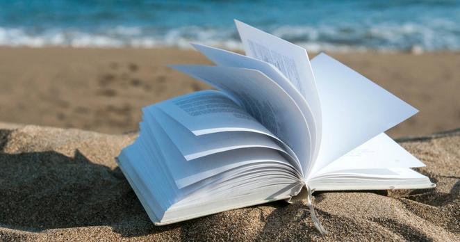 Ein Buch liegt auf einem Sandstrand, im Hintergrund sieht man das Meer.