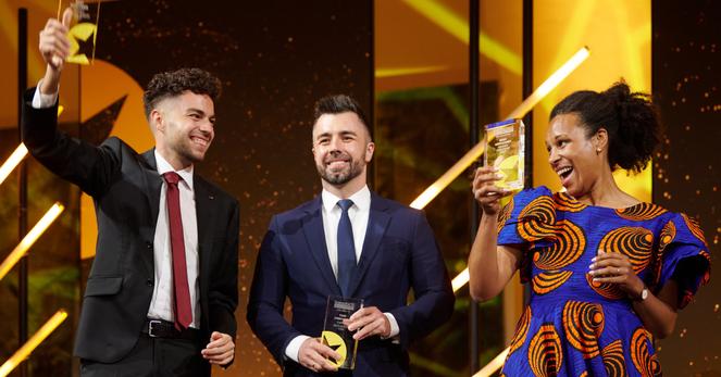 Nela Riehl, Member of the European Parliament for Volt, smiles joyfully while holding up her MEP Award on stage. She is wearing a vibrant blue and orange patterned dress. Two men in suits stand beside her, also holding awards and smiling, with a celebratory atmosphere and bright stage lighting in the background.