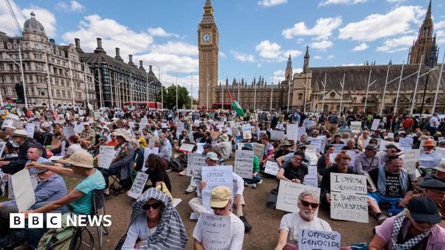 Hundreds of people hold signs reading 'I Oppose Genocide - I Support Palestine Action' while sitting in Parliament Square. Big Ben's clocktower is seen in the background as well as the Palace of Westminster.