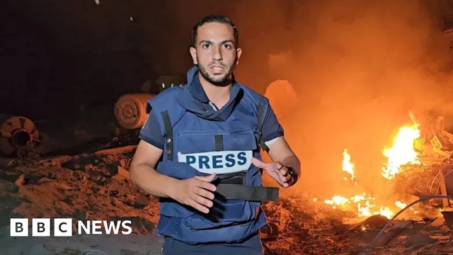 Anas al-Sharif is wearing a blue flak jacket with 'PRESS' written across the front, and he's standing in front of burning debris. Anas al-Sharif is wearing a blue flak jacket with 'PRESS' written across the front, and he's standing in front of burning debris.