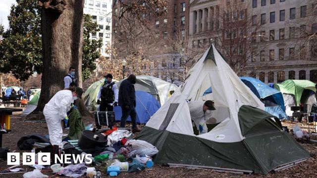 A tent encampment being removed from downtown Washington DC in 2023. Workers in hazmat suits wear face masks. Rubbish is seen strewn outside of one white and green tent. There are large trees and buildings visible outside of the edges of the public park